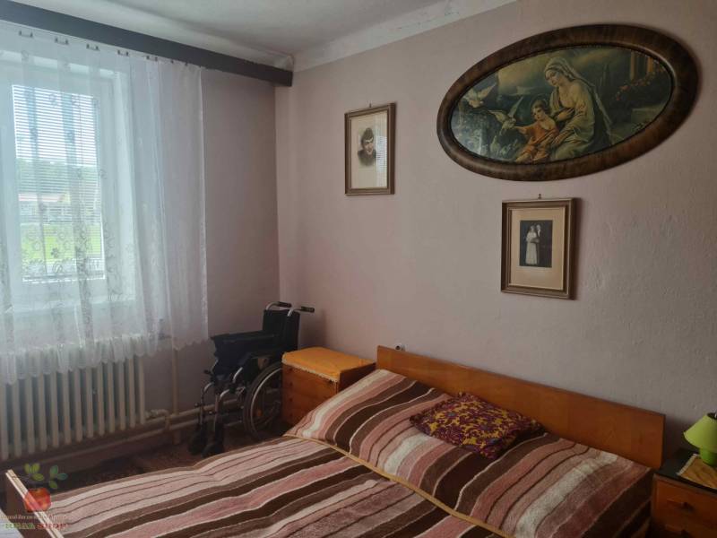 Spálňa v rodinnom dome s obrazmi, posteľou, invalidným vozíkom a oknom s čipkovými závesmi.
