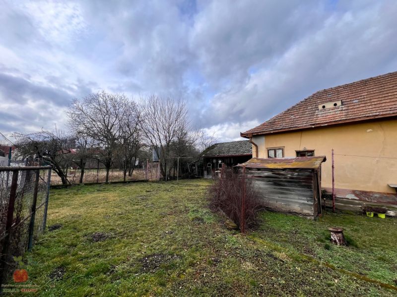 Dvor rodinného domu v Komjaticiach so staršou stavbou, trávnik a stromy pod oblačnou oblohou.